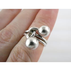 May include: A silver ring with a unique design. The ring features two silver spheres connected by curved bands. The ring is worn on a finger, showcasing its modern and minimalist style. The ring's polished surface reflects light, highlighting its metallic sheen.