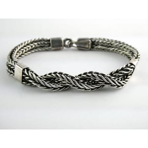 May include: A silver-toned braided bracelet with a secure clasp. The bracelet features a detailed woven design, with multiple strands intertwined to create a textured appearance. The metal has a polished finish, reflecting light. This piece of jewelry is suitable for everyday wear.