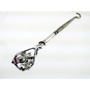 May include: A silver thimble with a purple glass stone and a thistle design. The thimble is attached to a long silver handle with a hook at the end.