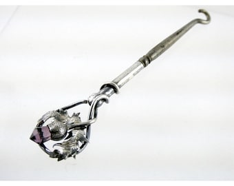 May include: A silver thimble with a purple glass stone and a thistle design. The thimble is attached to a long silver handle with a hook at the end.