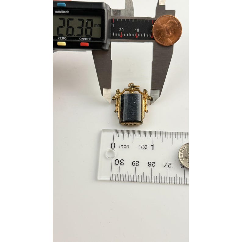 May include: A gold-coloured pendant with a rectangular, dark stone centre. The pendant is measured with a digital caliper, showing a measurement of 2.64 cm. A ruler and a penny are also in the image.
