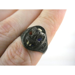 May include: A vintage silver-toned ring with a detailed design. The ring features a central emblem with red, white, and blue accents. The band has decorative cutouts. The word "SCOUT" is visible on the ring.