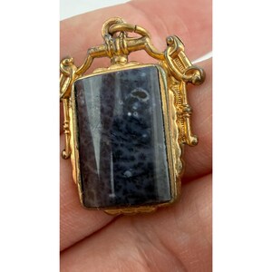 May include: A rectangular pendant with a gold-coloured frame and a dark blue and grey stone. The stone has a marbled appearance with white flecks. The frame has ornate detailing and a loop at the top for a chain.