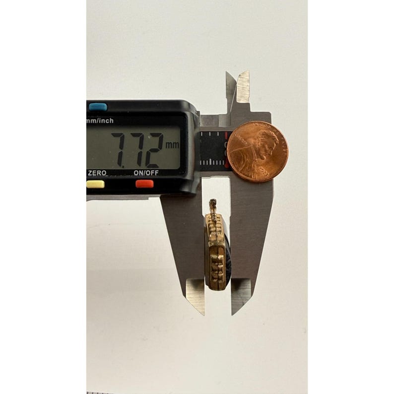 May include: A gold-coloured pendant is measured with a digital calliper, showing a measurement of 7.72 mm. A penny is placed next to the pendant for size comparison. The pendant has a dark blue centre and ornate gold detailing.