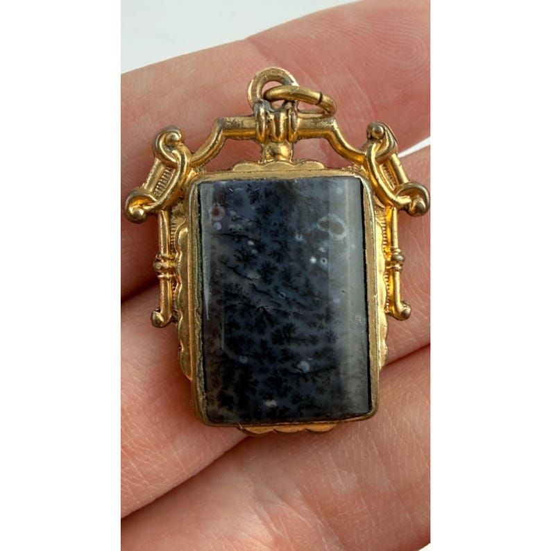 May include: A gold-coloured pendant featuring a rectangular, dark blue and white stone. The pendant has an ornate, filigree design around the stone and a small loop at the top for a chain. The stone has a unique, marbled pattern.