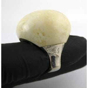 May include: A large, oval-shaped ring with a cream-colored stone set in a silver-toned metal band. The stone has subtle yellow and brown marbling. The ring's design is simple, with a focus on the prominent stone.