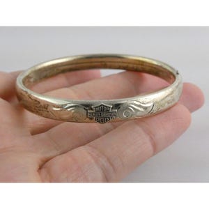Puede incluir: Un brazalete plateado con el logotipo de Harley-Davidson. El brazalete tiene un diseño grabado en los laterales y un cierre de bisagra. El logotipo de Harley-Davidson está centrado en el brazalete.