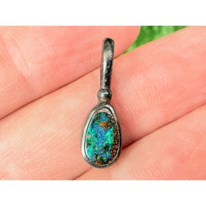 Pode incluir: Um pingente em forma de gota com uma armação de metal escuro e texturizado, apresentando uma incrustação iridescente vibrante com manchas cintilantes em tons de azul, verde e cobre. O pingente inclui uma argola de metal arredondada e robusta.