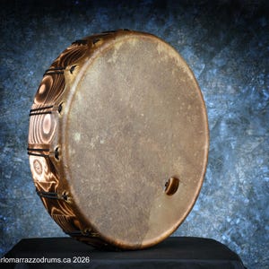 May include: A round, handcrafted drum with a natural hide head and a decorative, burnt wood frame. The drumhead is a light brown color, and the frame features a unique wood grain pattern. The drum has a small hole near the edge.