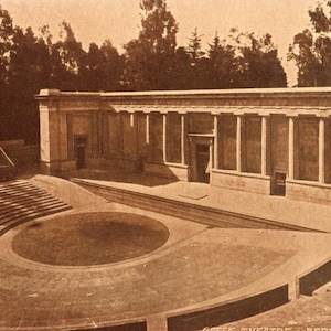 May include: Sepia-toned photograph of the Greek Theatre in Berkeley, California. The outdoor amphitheater features tiered seating, a stage area, and a colonnaded building. Trees are visible in the background.