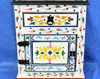 May include: White wooden nightstand with hand-painted floral designs in yellow, blue, pink, and green. The nightstand has a drawer and a door with black metal hinges and handles.