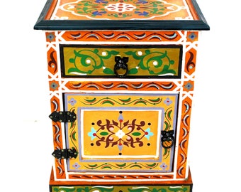 May include: A hand-painted wooden nightstand with a single drawer and a door. The nightstand is decorated with intricate floral patterns in shades of orange, yellow, green, blue, and white. The nightstand has black metal hardware.