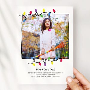 Peut inclure: Une carte de Noël avec une enfant tenant un râteau coloré. La carte a une bordure noire avec des guirlandes lumineuses et les mots "MERRY CHRISTMAS". Le message dit "Sending you the very best wishes for a joyful holiday season with love, Cayla, Gaby and Cory."