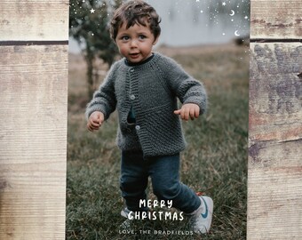 Puede incluir: Una tarjeta de Navidad con una foto de un niño pequeño que lleva un suéter de punto gris y pantalones vaqueros azules, de pie en un campo de hierba. La tarjeta tiene un fondo blanco con una dispersión de estrellas blancas y medias lunas. El texto "Feliz Navidad" y "Con cariño, Los Bradfields" está escrito en blanco en la tarjeta.