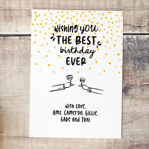 Op de afbeelding: Witte verjaardagskaart met de tekst "Wishing you THE BEST birthday EVER" in zwart schrift. De kaart heeft illustraties van twee handen die toosten met wijnglazen en gouden confetti. De kaart is ondertekend "With Love, Amy, Cameron, Gillie, Gabe And Toni."