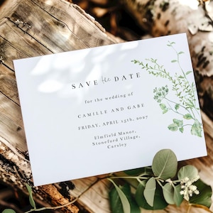 May include: Save the Date card with a watercolor floral design. The card is white with black text that reads "Save the Date" for the wedding of Camille and Gabe, Friday, April 15th, 2027. The venue is Elmfield Manor, Stoneford Village, Carsley.