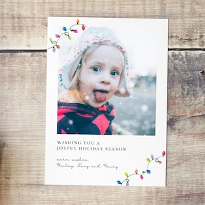 Puede incluir: Tarjeta de felicitaci&oacute;n navide&ntilde;a con la foto de un ni&ntilde;o sacando la lengua, con un dise&ntilde;o de luces de colores. La tarjeta incluye el texto "Wishing you a joyful holiday season" y los mejores deseos de Hailey, Lucy y Henry.