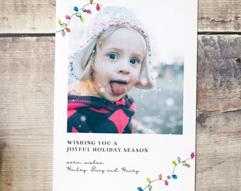 May include: Holiday greeting card featuring a child sticking out their tongue, with a festive string of lights design. The card includes the text "Wishing you a joyful holiday season" and warm wishes from Hailey, Lucy, and Henry.