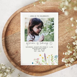 May include: A white invitation card with a photo of a young girl blowing on a dandelion. The text on the card reads "Join us to celebrate - Amani is turning 6. Saturday, August 8th from 3 - 6 PM. 525 Willow Street. RSVP to Allie. 123-456-789". The card is decorated with a watercolor illustration of wildflowers.