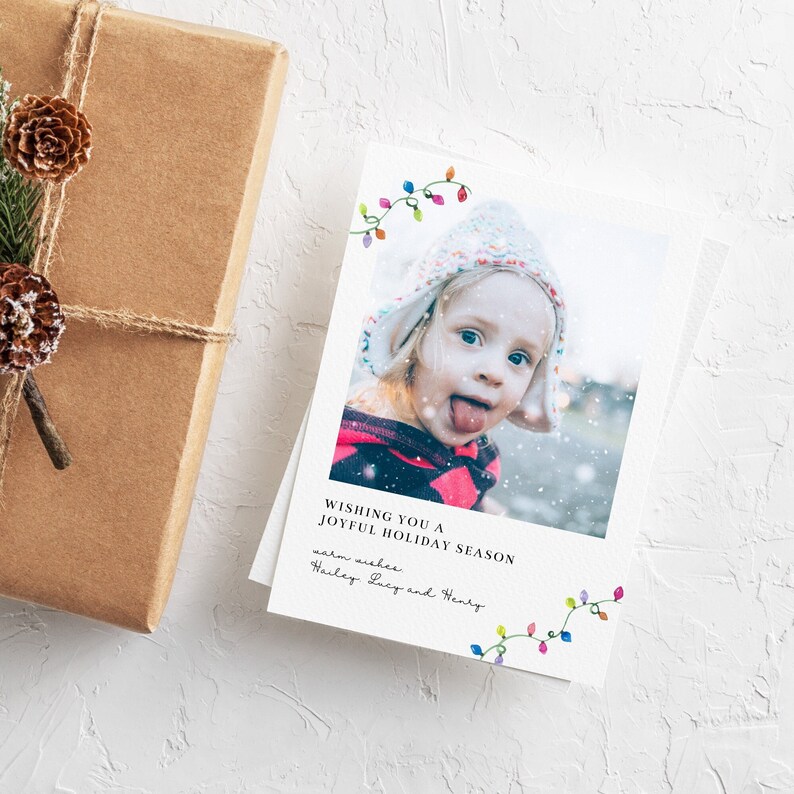 Puede incluir: Una tarjeta de felicitaci&oacute;n navide&ntilde;a con la foto de un ni&ntilde;o y el mensaje "Wishing you a Joyful Holiday Season". La tarjeta est&aacute; decorada con luces de colores y est&aacute; junto a un regalo envuelto con pi&ntilde;as.