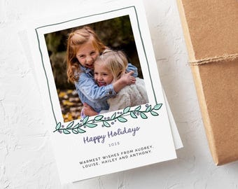 May include: A holiday greeting card featuring a photo of two young girls embracing. The card has a white background with a green and purple floral design and the text "Happy Holidays 2025". The card is next to a brown wrapped gift.