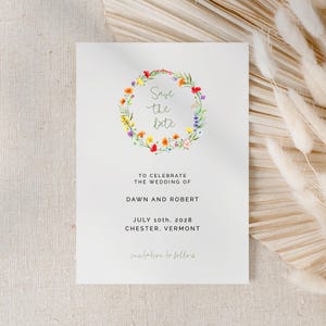 May include: Save the date wedding announcement with a floral wreath in shades of red, yellow, blue, and green. The text reads "Save the date" and includes the couple's names, wedding date, and location: July 10th, 2028, Chester, Vermont.