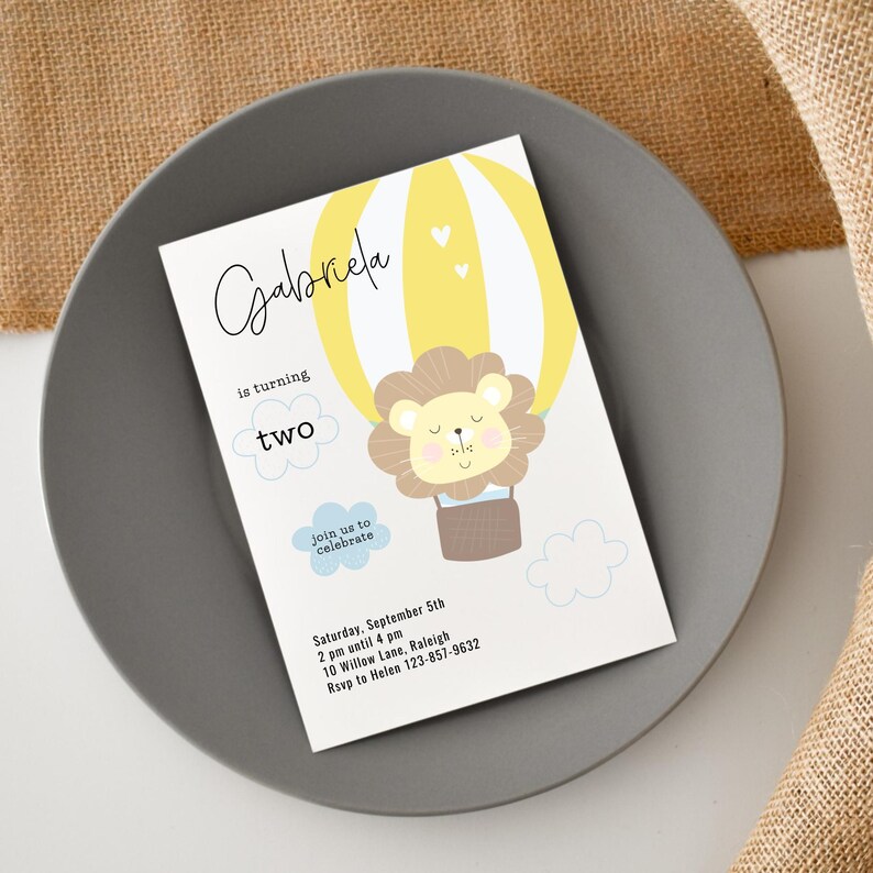 May include: A birthday invitation featuring a yellow hot air balloon with a lion illustration. The invitation reads "Gabriela is turning two" and includes event details. The invitation is on a gray plate.