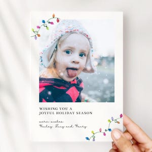 Puede incluir: Tarjeta de felicitaci&oacute;n navide&ntilde;a con una foto de un ni&ntilde;o sacando la lengua, con un gorro de punto y una parte superior rosa y negra. La tarjeta dice "WISHING YOU A JOYFUL HOLIDAY SEASON" y "warm wishes, Hailey, Lucy and Henry."
