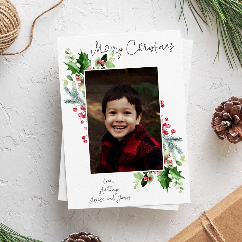 May include: A Christmas card with a watercolor border of holly and berries. A photo of a young boy smiling is in the center of the card. The text "Merry Christmas" is at the top of the card and "Love, Anthony, Louise and James" is at the bottom.