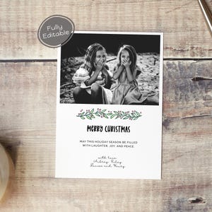 May include: A black and white photo of two girls covering their mouths with their hands. The photo is on a white card with a green and pink floral garland and the text "Merry Christmas". The card also includes the text "May this holiday season be filled with laughter, joy, and peace." and the names "Aubrey Riley, Lenise and Healy".
