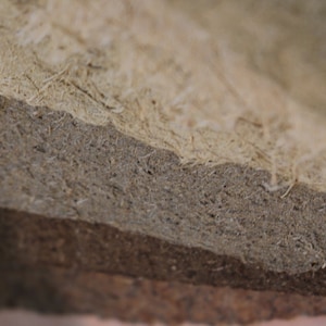 K&ouml;nnte beinhalten: Nahaufnahme eines geschichteten Materials mit nat&uuml;rlicher, strukturierter Optik. Die Schichten gehen von hellem Beige zu mittlerem Graubraun &uuml;ber, mit sichtbaren Fasern und einer rauen Oberfl&auml;che. Das Material scheint ein Verbundwerkstoff oder ein Naturprodukt zu sein.