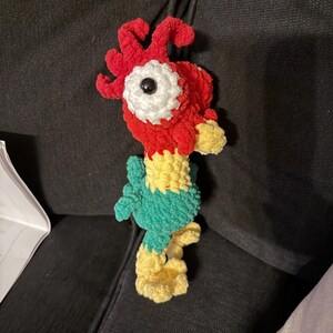 May include: A handmade crochet rooster toy. The rooster features a red head with a white and black eye, a yellow beak, and a green body. The legs and feet are yellow. The toy is made of soft, textured yarn.