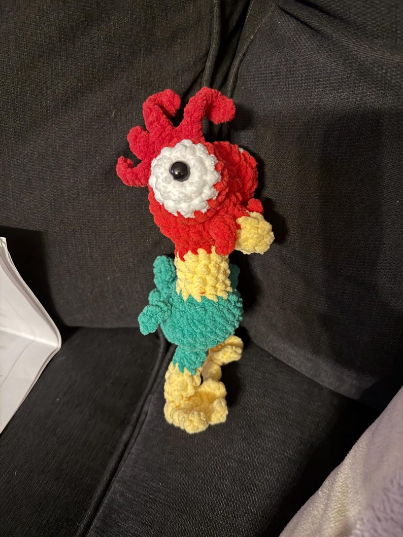 May include: A handmade crochet rooster toy. The rooster features a red head with a white and black eye, a yellow beak, and a green body. The legs and feet are yellow. The toy is made of soft, textured yarn.