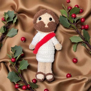 May include: A handmade crochet doll depicting a religious figure, wearing a white robe and a red sash. The doll has brown hair and a beard, with black eyes. The background is a golden fabric, decorated with holly branches and red berries.