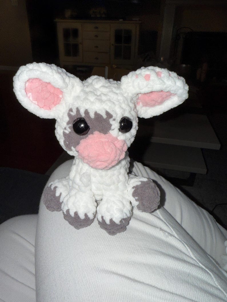 May include: A handmade crochet cow plushie. The cow is white with gray spots around the eyes and on the feet. The inner ears and nose are pink. The cow has large black eyes.