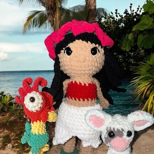 May include: Crocheted doll and two plush toys. The doll has black hair, a pink flower crown, and a red top. The toys include a red and yellow bird and a white lamb with pink ears. The scene is set on a beach with palm trees and ocean in the background.