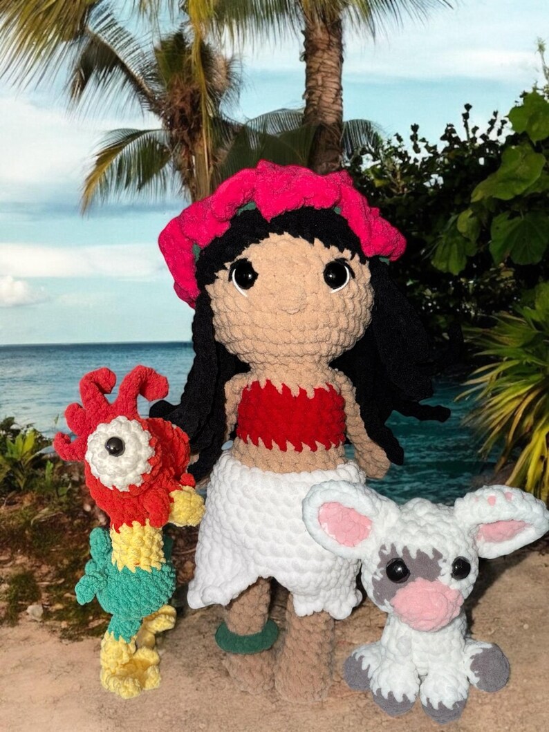 May include: Crocheted doll and two plush toys. The doll has black hair, a pink flower crown, and a red top. The toys include a red and yellow bird and a white lamb with pink ears. The scene is set on a beach with palm trees and ocean in the background.