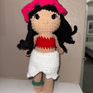 May include: A handmade crochet doll featuring a tan face, black hair, and large black eyes. The doll wears a red top, a white skirt, and a pink flower crown. Green bracelets adorn the ankles. The doll is approximately 10 inches tall.