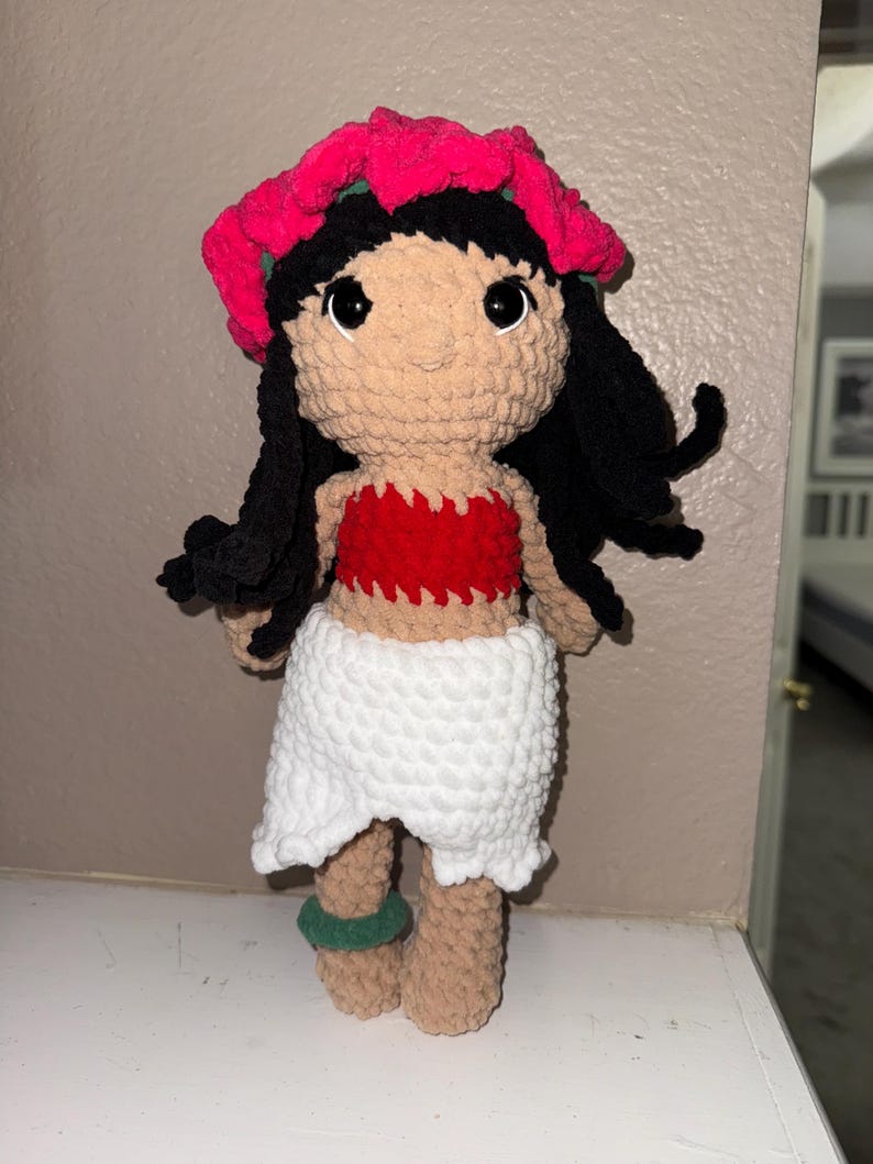May include: A handmade crochet doll featuring a tan face, black hair, and large black eyes. The doll wears a red top, a white skirt, and a pink flower crown. Green bracelets adorn the ankles. The doll is approximately 10 inches tall.