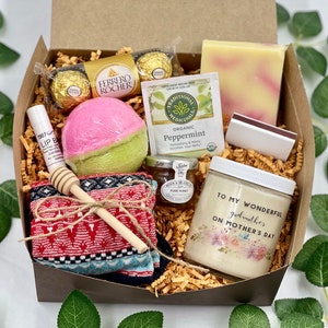 Puede incluir: Una caja de regalo llena de art&iacute;culos para una madrina, que incluyen una bomba de ba&ntilde;o, una vela, una barra de jab&oacute;n, un peque&ntilde;o frasco de miel, una caja de chocolates, una bolsita de t&eacute;, un b&aacute;lsamo labial, un par de calcetines y un cuchar&oacute;n de miel de madera. La caja est&aacute; forrada con virutas de papel marr&oacute;n. La vela tiene el texto "Para mi maravillosa madrina en el D&iacute;a de la Madre".