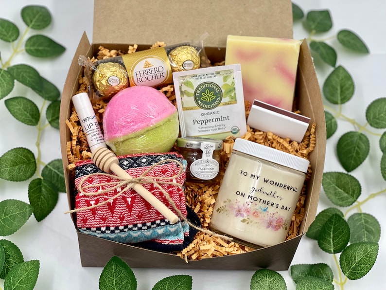 Puede incluir: Una caja de regalo llena de art&iacute;culos para una madrina, que incluyen una bomba de ba&ntilde;o, una vela, una barra de jab&oacute;n, un peque&ntilde;o frasco de miel, una caja de chocolates, una bolsita de t&eacute;, un b&aacute;lsamo labial, un par de calcetines y un cuchar&oacute;n de miel de madera. La caja est&aacute; forrada con virutas de papel marr&oacute;n. La vela tiene el texto "Para mi maravillosa madrina en el D&iacute;a de la Madre".