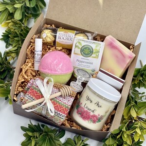 May include: A brown gift box filled with a variety of self-care items, including a bath bomb, soap, a candle, honey, and chocolates. The candle has the text "You Are Loved" on the label.