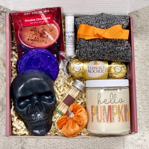 May include: A pink gift box filled with fall-themed items, including a black skull soap, a purple bath bomb, a candle that says "hello pumpkin", a package of chocolate candies, a pair of gray socks, and a hot cocoa mix packet.