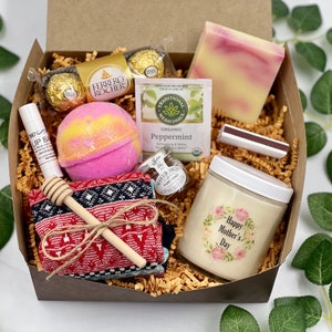 Op de afbeelding: Een bruine cadeaudoos gevuld met verschillende items, waaronder een badbom, een kaars, een zeep, chocolade en een paar sokken. De kaars heeft een bloemenmotief en de tekst "Gelukkige Moederdag".