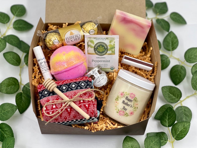 Op de afbeelding: Een bruine cadeaudoos gevuld met verschillende items, waaronder een badbom, een kaars, een zeep, chocolade en een paar sokken. De kaars heeft een bloemenmotief en de tekst "Gelukkige Moederdag".