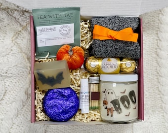 May include: A pink gift box filled with Halloween themed items, including a ghost candle, a purple bath bomb, a bar of soap with a bat design, a box of chocolates, a tea bag, a pumpkin, a pair of socks, and a lip balm.