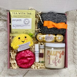 May include: A pink gift box filled with a variety of items, including a crocheted lemon, tea, socks, chocolates, a lip balm, matches, and a candle that says "nurse life".