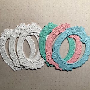 May include: Set of six oval-shaped picture frames with ornate detailing. Three frames are white, one is light blue, one is light pink, and one is light green.