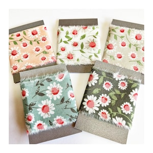 May include: Four samples of floral patterned fabric ribbon on brown card stock. The patterns feature white daisies with red centers on a variety of backgrounds including peach, cream, green, and dark green.