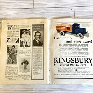 Peut inclure: Un camion jouet bleu et rouge avec une benne en bois, portant l'inscription "Kingsbury Motor Driven Toys". Le camion est charg&eacute; de bo&icirc;tes et poss&egrave;de un volant, un levier et un moteur. La l&eacute;gende indique "Chargez-le et partez !" et "Kingsbury Motor Driven Toys".